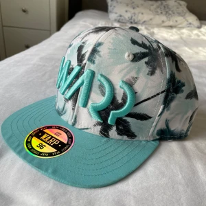 Turkos keps med palmmönster från WARP - Snygg keps från WARP med turkos skärm och broderad logga. Kepsen har ett tropiskt palmmönster i svart och vitt, perfekt för sommaren. Justerbar passform baktill.