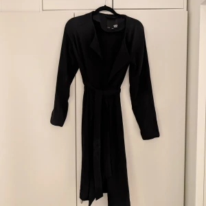 Slutsåld! Svart trenchcoat från Lager 157 - Snygg svart trenchcoat från Lager 157 i 100% polyester. Den har långa ärmar och ett stilrent bälte i midjan för en smickrande passform. Slutsåld modell som inte längre säljs i affär! Perfekt för en sofistikerad look.