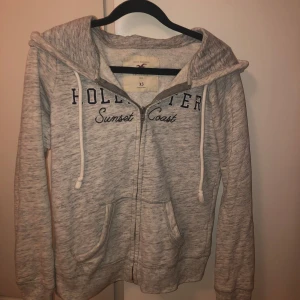 Grå hoodie från Hollister - Säljer en grå hoodie från Hollister med texten 'Sunset Coast' på bröstet. Den har en dragkedja framtill och justerbara snören. Säljer pågrund av inte kommer till användning. Skulle säga att den är i mycket bra skick ❤️🫰