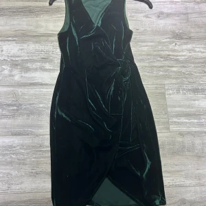 Mörkgrön sammet klänning - Elegant mörkgrön klänning i sammet med omlott-design och knytning i midjan. Klänningen är ärmlös och har en v-ringad halslinje, vilket ger en stilren look. Använd bara en gång under Julafton, är i mycket bra skick!