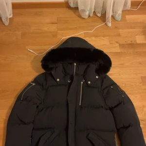 Moose knuckles  - Snygg svart dunjacka med pälskrage och dragkedja. Perfekt för kalla dagar med sin varma och stilrena design. Jackan har flera praktiska fickor och en justerbar huva.