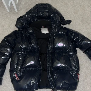 Svart pufferjacka från Tommy Hilfiger - Snygg svart pufferjacka från Tommy Hilfiger med glansig finish och broderad logga på ärmen. Jackan har en praktisk huva och dragkedja framtill. Perfekt för kyliga dagar.
