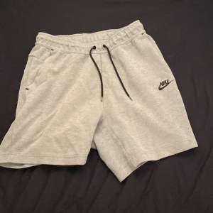 Gråa Nike tech shorts  - Säljer ett par grå Nike shorts med snörning i midjan och sidofickor. Perfekta för en avslappnad stil eller träning. Klassisk Nike-logga på benet.