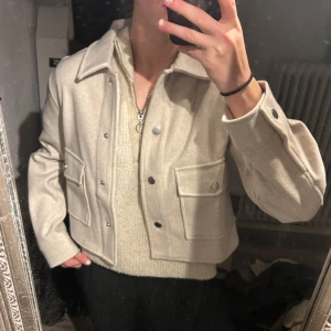 Beige jacka från Stradivarius - Snygg beige jacka från Stradivarius i ullblandning. Jackan har en klassisk krage, knappar framtill och två stora fickor. Perfekt för höst och vår med sin stilrena design. En kortare ”kappa” som är oanvänd, lappen sitter kvar. (Är 170 lång)
