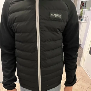 Svart pufferjacka från McKenzie - Snygg svart pufferjacka från McKenzie med dragkedja framtill. Jackan har långa ärmar och en huva för extra skydd mot väder och vind. Perfekt för kyligare dagar. Den har även vadd i luvan och inne i armarna så den håller värmen bra. Den är helt ny, har aldrig används så mycket bra skick!  