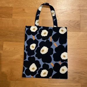 Marimekko Unikko hemmastadd shoppingväska i bomull - Snygg shoppingväska med stort blommönster i svart och beige på en blå bakgrund. Väskan har två handtag och är tillverkad i bomull. Perfekt för att bära med sig det nödvändigaste.
