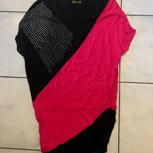 Svart och rosa topp med strassdetaljer - Trendig svart och rosa topp med korta ärmar och strassdetaljer på ena axeln. Toppen har en asymmetrisk design med en kombination av färger som ger en unik look.