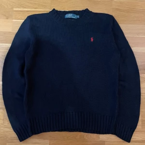 Marinblå stickad tröja från Polo Ralph Lauren - Säljer en klassisk marinblå stickad tröja från Polo Ralph Lauren med det ikoniska röda broderade logotypen på bröstet. Tröjan är tillverkad i 100% bomull och har ribbstickade muddar vid ärmslut och nederkant. Perfekt för en stilren look.
