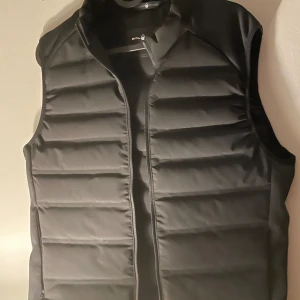 Svart dunväst från Sail Racing  - Snygg och funktionell svart dunväst från Sail Racing. Perfekt för kyliga dagar med sin quiltade design och dragkedja framtill. Västen är tillverkad i polyester och dun, vilket ger både värme och stil. 