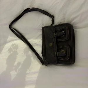 Mörkbrun axel/ handväska med spännen - Snygg mörkbrun axel/handväska i skinn med justerbar axelrem. Väskan har två framfickor med spännen och en dragkedja på baksidan för extra förvaring. Perfekt för att hålla dina saker organiserade och säkra.