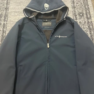 Marinblå vindjacka från Sail Racing - Snygg marinblå vindjacka från Sail Racing med dragkedja och huva. Perfekt för blåsiga dagar. Jackan har en stilren design med logotyp på bröstet.
