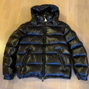 Moncler maya - Snygg svart pufferjacka med glansig yta och praktisk huva. Jackan har dragkedja framtill och två fickor med dragkedjor. Perfekt för kyliga dagar. Pris går att diskutera!