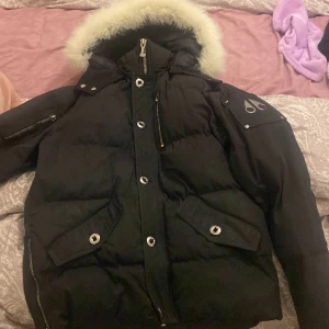 Svart dunjacka från Moose Knuckles - Säljer en svart dunjacka från Moose Knuckles i mycket bra skick. Jackan har en fluffig pälsfodrad huva och snygga detaljer som dragkedja och knappar. Perfekt för kalla vinterdagar! 🖤