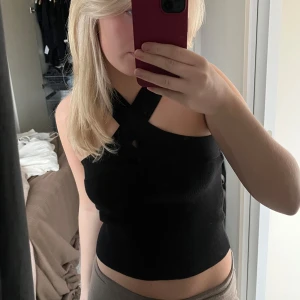 Svart ribbad topp - Säljer en snygg svart ribbad topp, den har en korsad detalj vid halsen och är ärmlös. 