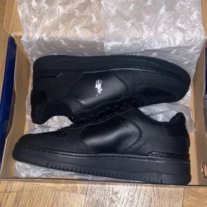 Svarta sneakers i skinn - Säljer ett par svarta sneakers i skinn med en cool logga på sidan. De är helt nya och kommer i originalkartong. Perfekta för både vardag och fest! Skorna har en klassisk design med snörning och en bekväm passform.