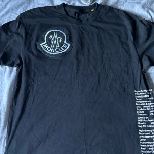Svart t-shirt från Moncler - Säljer en svart t-shirt från Moncler i mycket bra skick. Den har ett stort Moncler-märke på bröstet och textdetaljer på baksidan. Perfekt för en stilren och avslappnad look. Passar bra till både vardag och fest!