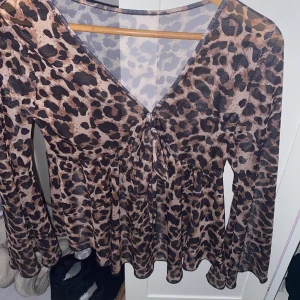 Leopardmönstrad topp med knytning - Säljer en snygg leopardmönstrad topp med v-ringning och knytning framtill. Toppen har långa ärmar och är perfekt för en utekväll eller fest. Materialet är lätt och luftigt, vilket gör den bekväm att bära. Aldrig använd och märket är okänt. Ser stor ut men passar mycket bättre på