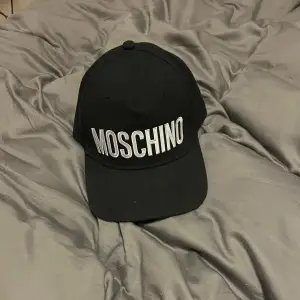 Snygg svart keps från Moschino med broderad logga i vitt framtill. Kepsen har en justerbar rem baktill för perfekt passform. Perfekt för både vardag och festliga tillfällen. En stilren accessoar som passar till det mesta!