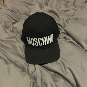Svart keps från Moschino - Snygg svart keps från Moschino med broderad logga i vitt framtill. Kepsen har en justerbar rem baktill för perfekt passform. Perfekt för både vardag och festliga tillfällen. En stilren accessoar som passar till det mesta!