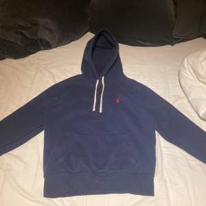 Säljer en snygg blå hoodie från Ralph Lauren i nyskick. Den har en klassisk design med en liten röd logga på bröstet och vita snören i huvan. Perfekt för en avslappnad stil och passar till de flesta tillfällen. Superbekväm och stilren! Nypris ligger på 2000kr