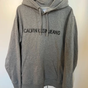 Grå hoodie från Calvin Klein Jeans - Säljer en snygg och bekväm grå hoodie från Calvin Klein Jeans. Perfekt för både vardag och chill. Den har en klassisk passform med känguruficka och justerbar huva. Texten 'CALVIN KLEIN JEANS' pryder framsidan. Passar perfekt till jeans eller joggers! Pris kan diskuteras.