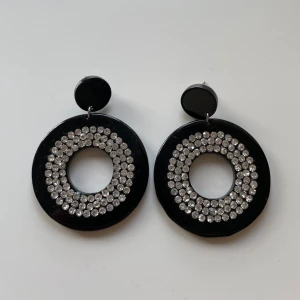 Svarta örhängen med glittriga stenar - Snygga svarta örhängen i cirkelform med glittriga stenar. Perfekta för en festlig kväll eller när du vill lägga till lite bling till din outfit. De har en modern design med en stor cirkel och mindre cirkel upptill. Ett måste för smyckesamlingen!