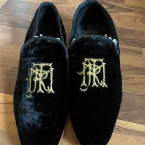 Svarta loafers i sammet från Asos - Snygga svarta loafers i sammet från Asos med guldbroderi på ovansidan. Perfekta för en festlig kväll eller en stilren look. Skorna är i bra skick och har en elegant design som verkligen sticker ut.