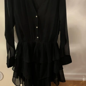 Svart klänning med volanger - Säljer en elegant svart klänning med volanger och knappar framtill. Den har långa ärmar och en v-ringad design som ger en stilren look. Perfekt för festliga tillfällen eller en utekväll. Materialet är lätt och luftigt, vilket gör den bekväm att bära. 