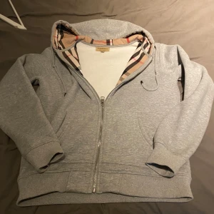 Grå hoodie från Burberry - Säljer en snygg grå hoodie från Burberry i mycket bra skick. Den har en klassisk dragkedja och den ikoniska Burberry-rutiga insidan av huvan. Perfekt för höst och vår, med en broderad logga på bröstet. Superbekväm och stilren!