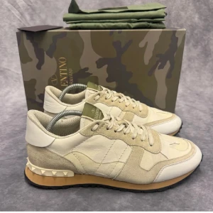 Valentino Rockrunner i beige och vit - Säljer ett par snygga Valentino Rockrunner sneakers i beige och vit. De har en cool camo-låda och är i nytt skick. Skorna har snörning och är gjorda av en mix av skinn och mocka. Perfekta för både vardag och fest!