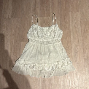 Vit spetstopp med volangdetaljer - Söt vit topp med tunna axelband och vacker spets upptill. Kjolen är luftig med volangdetaljer som ger en romantisk känsla. Perfekt för sommarens fester eller en dag på stranden. Topp är i mycket bra skick och passar perfekt för en bohemisk look.