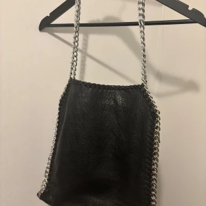 Tiamo svart axelväska med kedjedetaljer - Snygg svart axelväska i skinn från Tiamo med coola kedjedetaljer längs kanterna. Väskan har en praktisk dragkedja både inuti och utanpå, perfekt för att hålla ordning på dina saker