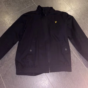 Lyle and Scott Jacka - Säljer en stilren mörkblå jacka från Lyle & Scott. Den har en dragkedja framtill och två knappar på fickorna. Perfekt för höst, varmare vinter och vår. Passar både till vardags och lite finare tillfällen.