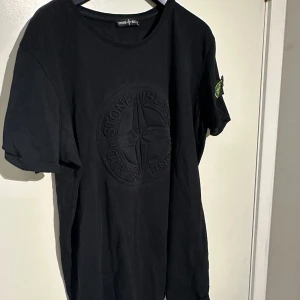 Svart t-shirt från Stone Island - Säljer en svart t-shirt från Stone Island i storlek L. Den har en diskret broderad logga på bröstet och den ikoniska Stone Island-märket på ärmen. Perfekt för en stilren och muscle fit look. T-shirten är i bra skick och gjord av mjuk bomull.