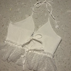 Vit topp från Chiquelle - Säljer en superfin vit topp från Chiquelle i storlek medium. Den har en söt knytning i nacken och en volangdetalj nedtill. Perfekt för sommardagar eller en festlig kväll. Toppen är i ett ribbat material med prickigt mönster på volangen.