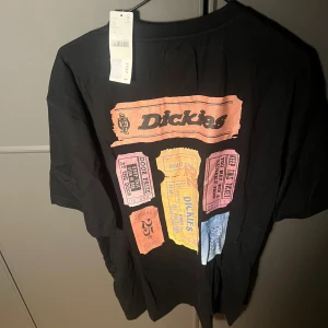 Svart t-shirt från Dickies - Säljer en svart t-shirt från Dickies i storlek L. Den har ett coolt tryck med olika färgglada etiketter på baksidan och en liten logga på framsidan. Perfekt för vardagsbruk eller avslappnade tillfällen. Helt ny med prislapp kvar!