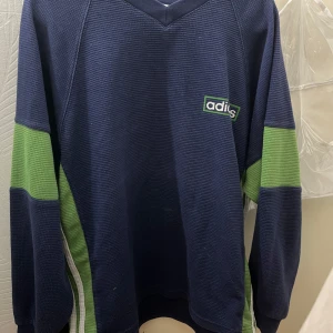 Blå och grön tröja från Adidas - Säljer en snygg blå tröja från Adidas med gröna detaljer. Tröjan har en v-ringad hals och långa ärmar. Perfekt för en sportig look eller en avslappnad dag. Den är i bra skick och redo för nya äventyr!