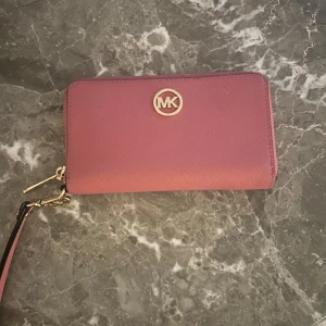 Rosa plånbok från Michael Kors - Säljer en snygg rosa plånbok från Michael Kors i mycket bra skick. Den har en praktisk dragkedja och en avtagbar handledsrem. Perfekt för att hålla ordning på kort och kontanter med stil!