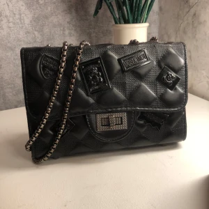 Svart quiltad axelväska med kedja - Snygg svart quiltad axelväska med coola detaljer och en kedjerem. Den är mochino inspirerad och är i ett bra skick, dock så har färgen på kedjan börjat släppa lite men inget man lägger märke till.