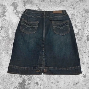 Mörkblå jeanskjol - Säljer en snygg mörkblå jeanskjol i bra skick. Kjolen har en klassisk femficksdesign med dekorativa sömmar på bakfickorna och en knappdetalj framtill. Perfekt för både vardag och festliga tillfällen. Passar bra till en avslappnad stil med sneakers eller klä upp med klackar.                       Måtten följer: Midja: 80cm Total längd: 56 cm