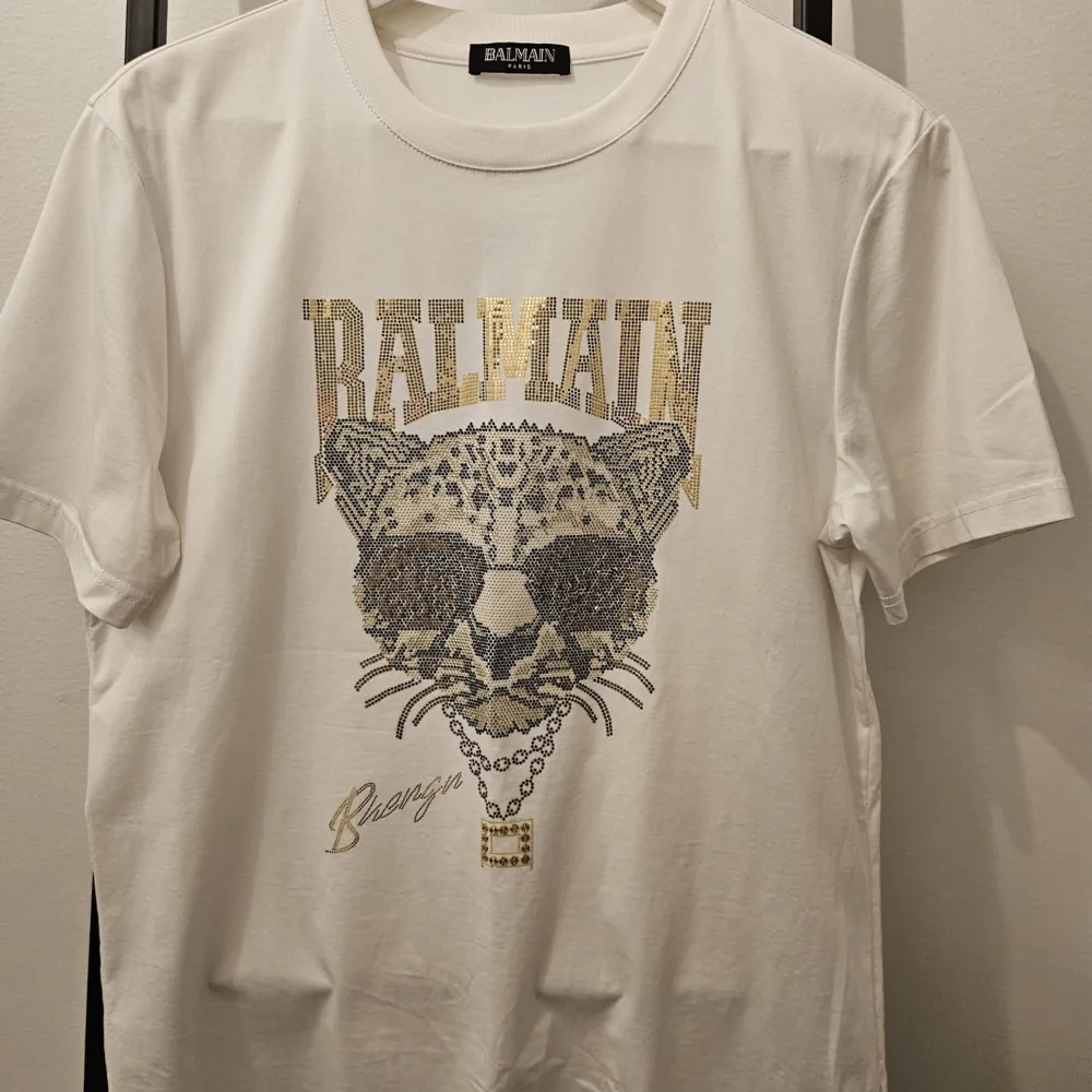 Säljer en snygg vit t-shirt från Balmain i storlek M. Den har ett coolt leopardmotiv med solglasögon och texten 'Balmain' i glittriga detaljer. Perfekt för en avslappnad men stilren look. Passar bra till både jeans och shorts!. T-shirts.