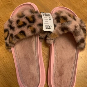 Rosa tofflor med leopardmönster - Supermysiga rosa tofflor i storlek 37 med fluffig leopardmönstrad rem. Perfekta för att hålla fötterna varma och stiliga hemma. Sulan är också rosa och har ett vågigt mönster för extra grepp. De är i nytt skick och redo att användas direkt!