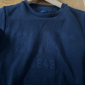 Mörkblå sweatshirt från GANT - Säljer en snygg mörkblå sweatshirt från GANT i bra skick. Den har ett diskret broderat GANT 1949-märke på framsidan. Perfekt för höst och vinter med sin bekväma passform och långa ärmar. Passar både till vardags och lite mer avslappnade tillfällen. Den är för liten för mig där av säljer jag den