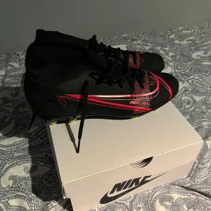 Nike Mercurial fotbollsskor - Säljer ett par svarta Nike Mercurial fotbollsskor i nytt skick. De har en snygg röd detaljdesign och är perfekta för fotbollsspel på gräs. Skorna har snörning och en tight passform för bästa kontroll och komfort på planen. Storlek 42