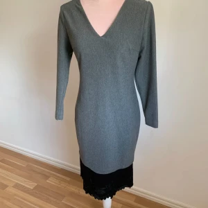 Grå klänning med spetsdetaljer - Elegant grå klänning med långärmad design och v-ringning. Baksidan har en unik öppen rygg med dragkedja och rosa inslag. Nederdelen pryds av svarta spetsdetaljer som ger en sofistikerad touch.