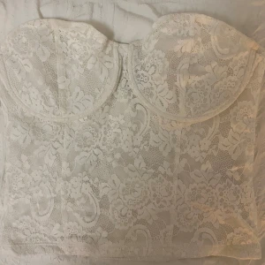 Vit spetskorsett från Gina Tricot - Säljer en elegant vit spetskorsett från Gina Tricot. Den har en vacker blommig spetsdesign och är perfekt för att ge en romantisk touch till din outfit. Korsetten har en tight passform och är axelbandslös.
