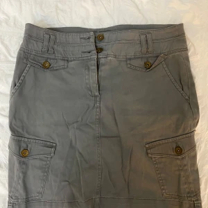 Grå kjol med knappar - Snygg cargo grå  kjol med knappar framtill och praktiska fickor. Kjolen har en klassisk design med bälteshällor . Perfekt för en stilren look.