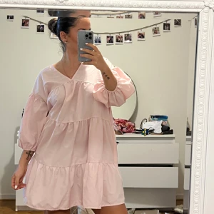 Ljusrosa klänning med volanger - Söt ljusrosa klänning med volanger och v-ringad hals. Den har trekvartsärmar och en luftig passform som ger en avslappnad look. Perfekt för varma dagar och ger en romantisk touch till din stil.