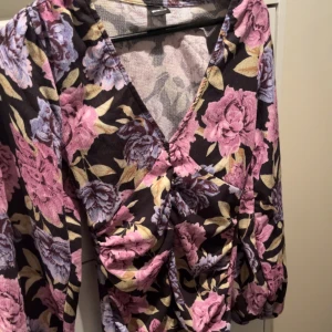 Blommig klänning med v-ringning - Säljer en vacker blommig klänning med v-ringning och trekvartsärmar. Klänningen har ett färgglatt mönster i rosa, lila och grönt på en mörk bakgrund. Den är ifrån ginatricot och helt oanvänd.