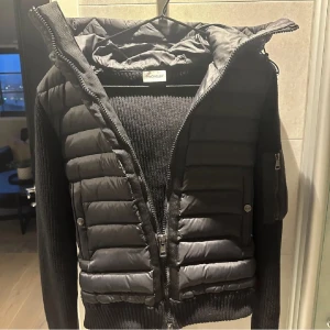 Moncler cardigan  - Snygg svart   Moncler cardigan med quiltad design och dragkedja. Jackan har långa ärmar och en praktisk huva. Perfekt för kyligare dagar med stil.